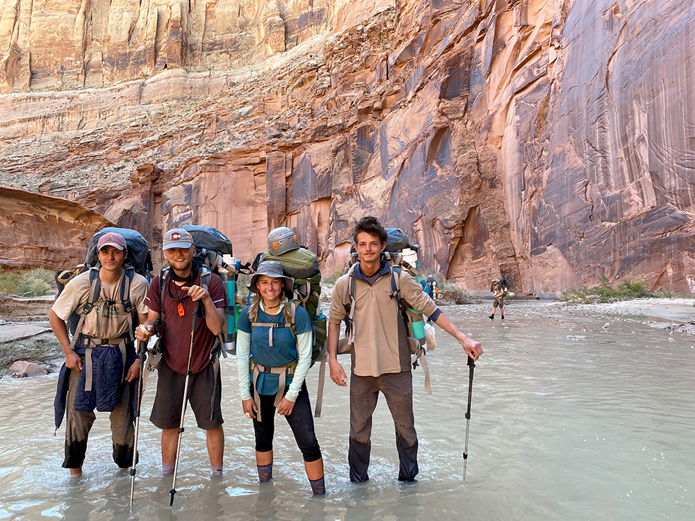 A group of people stand in a shallow river in a red desert canyon.